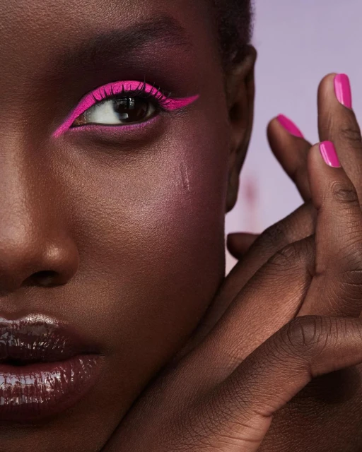Close-up of a dark-skinned woman with bright pink graphic eyeliner and glossy lips, set against a blurred soft lavender background.
