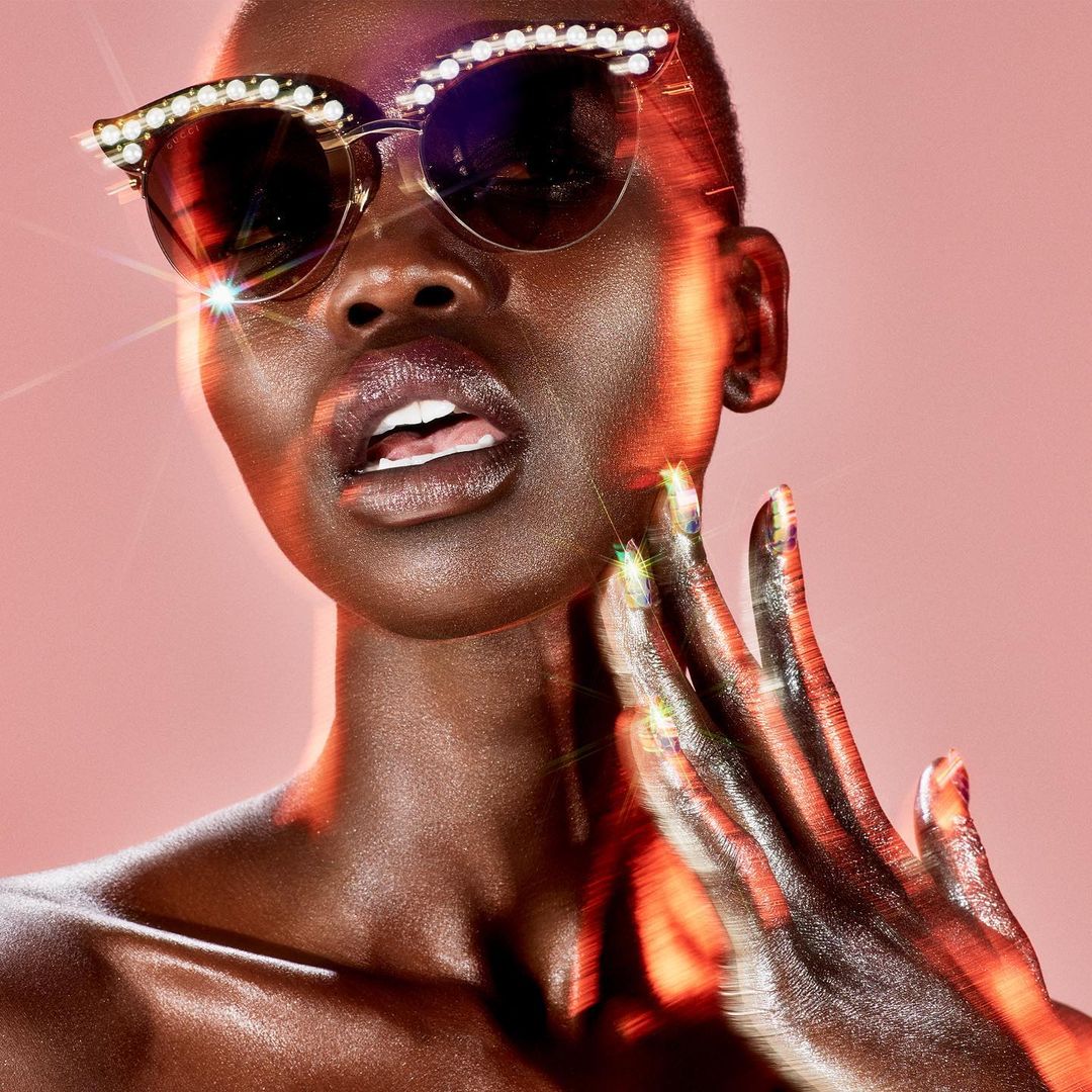 Close-up of a fashionable adult Black female wearing sunglasses with pearl embellishments, with her hand near her face, against pink backdrop.