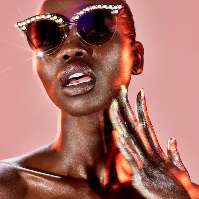 Close-up of a fashionable adult Black female wearing sunglasses with pearl embellishments, with her hand near her face, against pink backdrop.