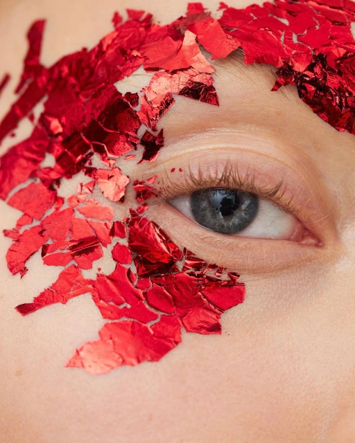 Close-up of a woman's face featuring one blue eye, with red shards affixed around the eye and brow, studio lighting.