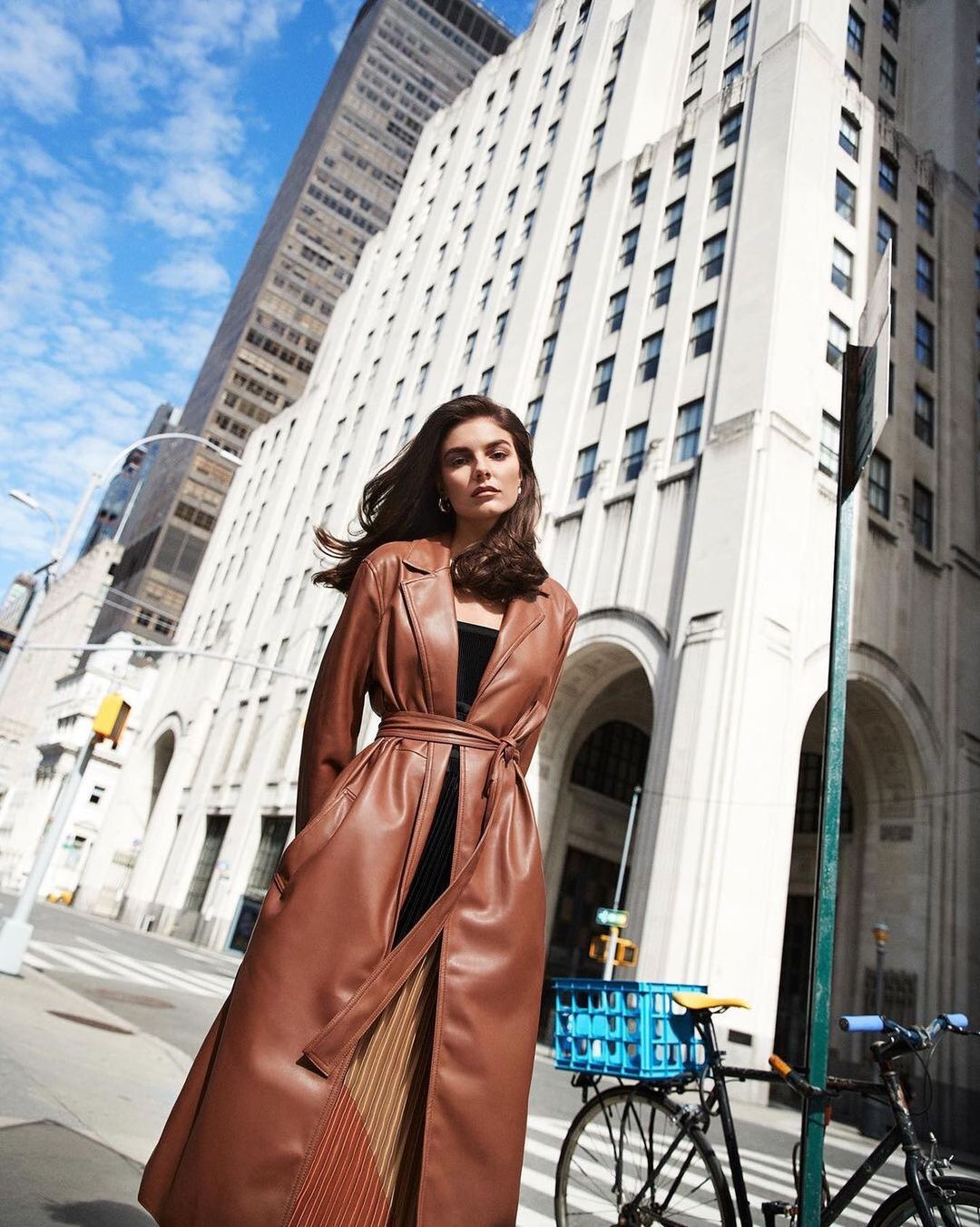Brunette woman in long brown coat poses in city street with tall buildings and bicycle. Sunny day, low angle, fashion editorial.