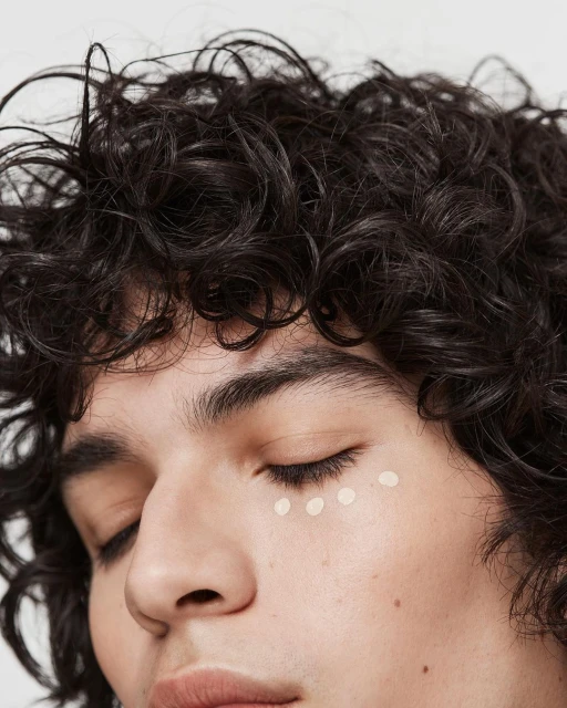 Close-up of a person with closed eyes and dark curly hair, with three spots of concealer under their eye in a well-lit indoor shot.