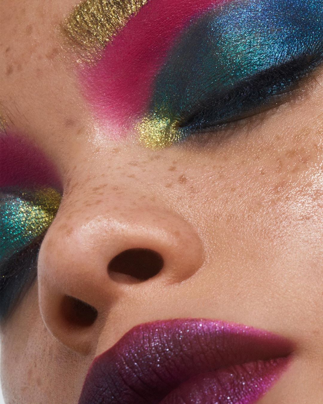 Close-up of a model wearing glittery gold, pink, and blue eyeshadow along with dark, sparkling lipstick in a beauty and makeup concept.