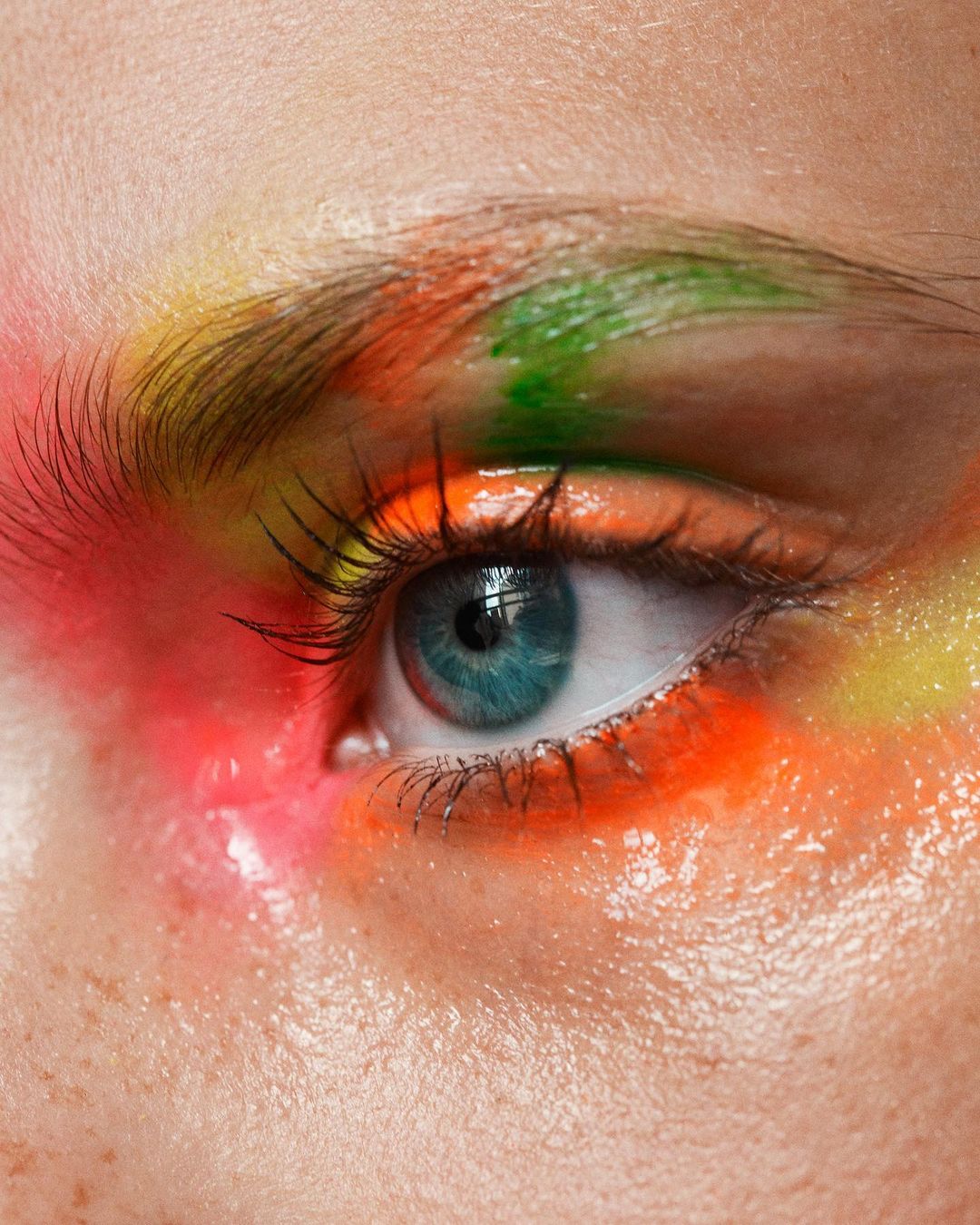 Close-up of a human eye with vibrant makeup featuring orange, yellow, and green colors around the eye and eyebrow.