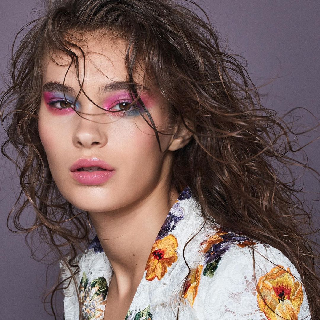 Close-up of a young woman with curly brown hair and striking pink and blue eyeshadow, wearing a white floral jacket against a grey background.