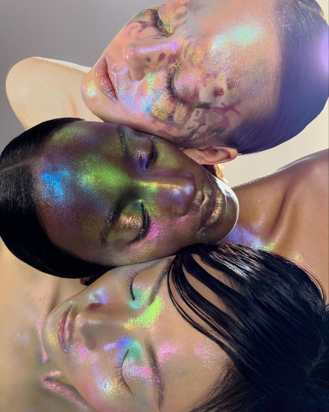 Close-up beauty shot featuring three female faces stacked vertically, faces covered in iridescent makeup that reflects green, blue and pink.