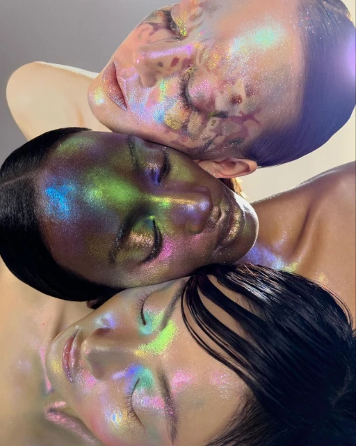 Close-up beauty shot featuring three female faces stacked vertically, faces covered in iridescent makeup that reflects green, blue and pink.