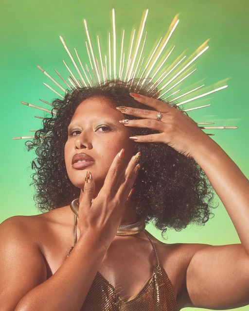 Portrait of a woman with dark curly hair wearing a golden headpiece and gold jewelry against a gradient green background in studio lighting.