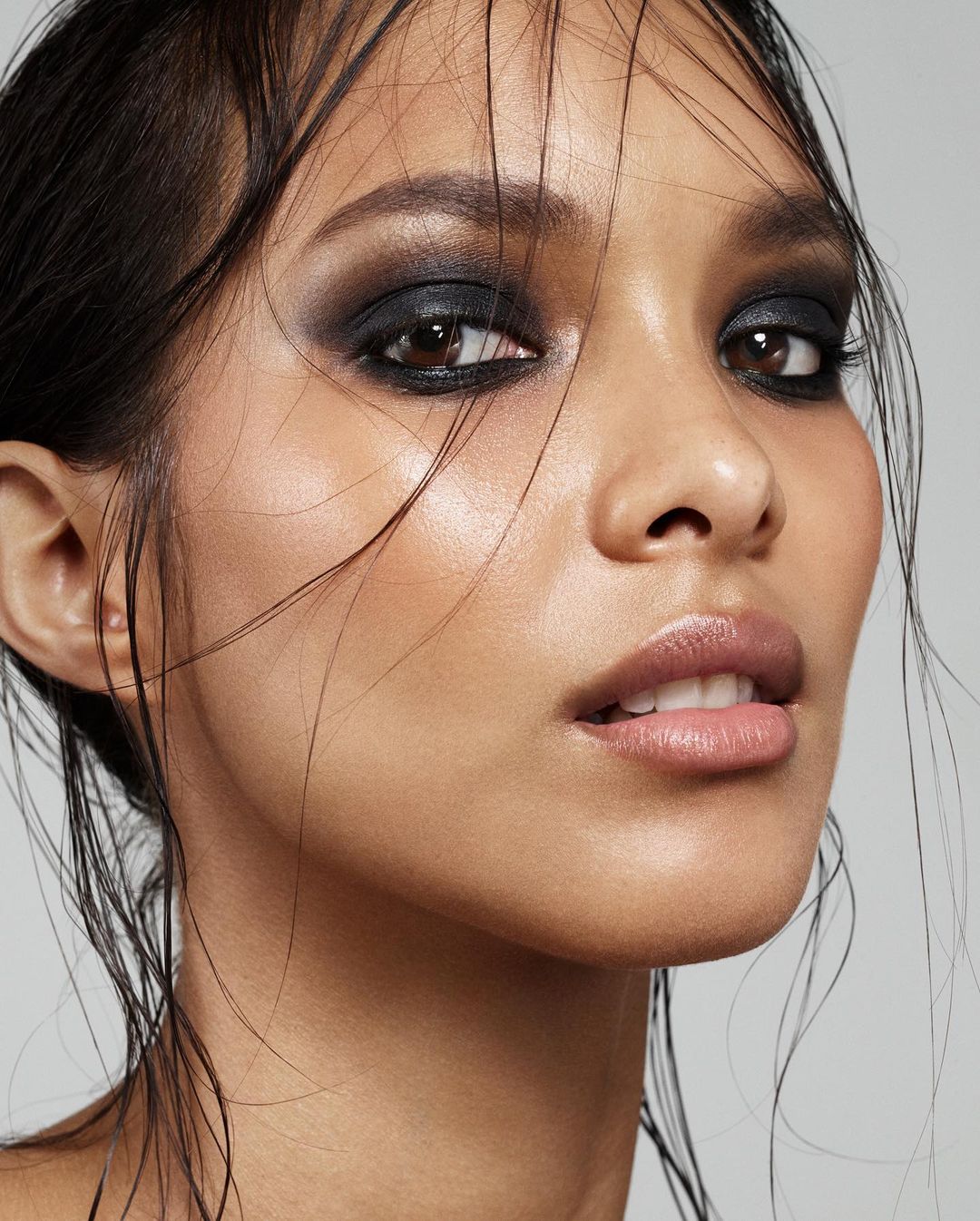 Close-up portrait of a young adult woman with wet dark hair and dark smokey eyeshadow, posing indoors against a neutral grey background.