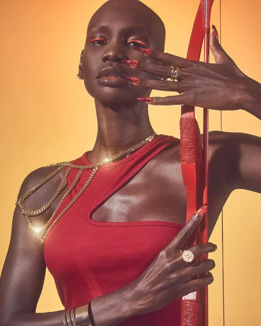 Striking portrait of an adult female with a shaved head in a red dress, holding a red bow, set against a vibrant orange backdrop.