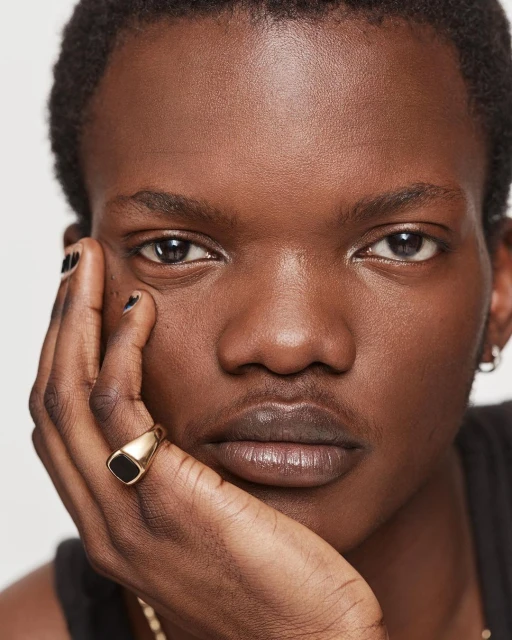 Close-up studio portrait of a dark-skinned adult man with black fingernails and wearing a gold ring, gazing directly at the camera.