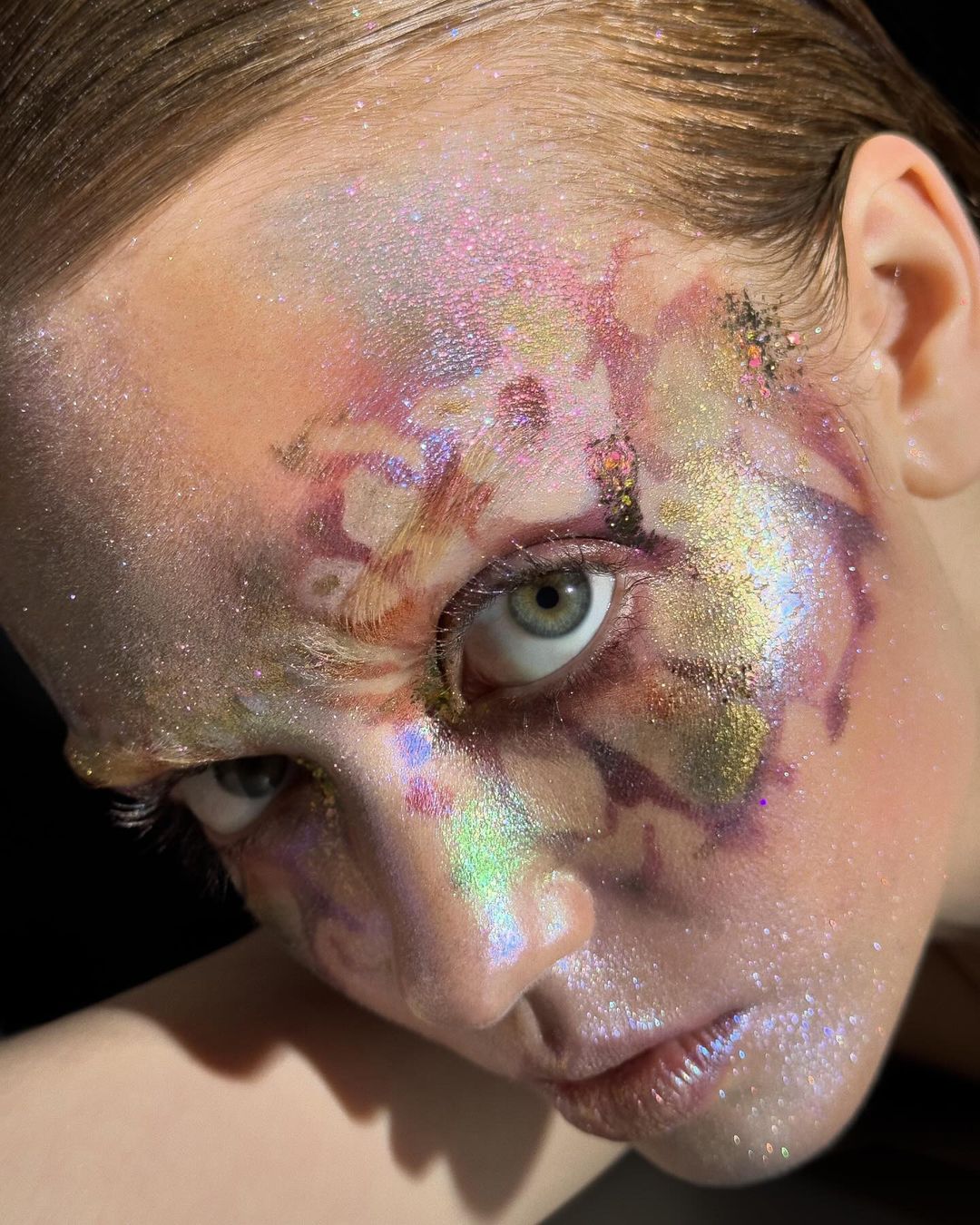 Close-up portrait of a model with colorful glitter makeup on her face and shoulders against a dark background, showcasing beauty and artistry.