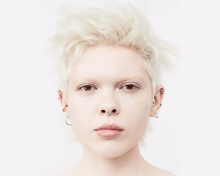 Close-up studio portrait of a fair-skinned adult woman with short white hair, facial and ear piercings, against a solid white backdrop.