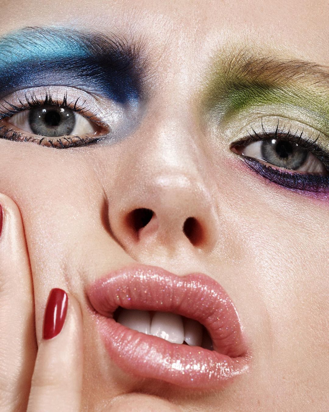 Close-up of a woman's face with colorful eyeshadow in blue, green, and purple, shiny pink lipstick, and red nail polish, beauty shot.