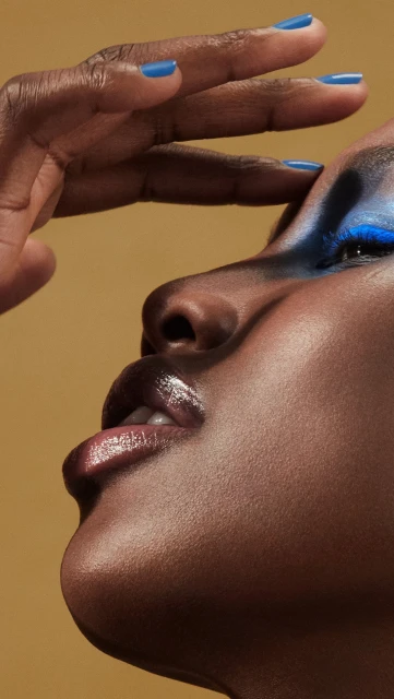 Close-up portrait of a Black woman with vibrant blue eyeshadow and nail polish, her hand raised to her forehead in a studio shot.