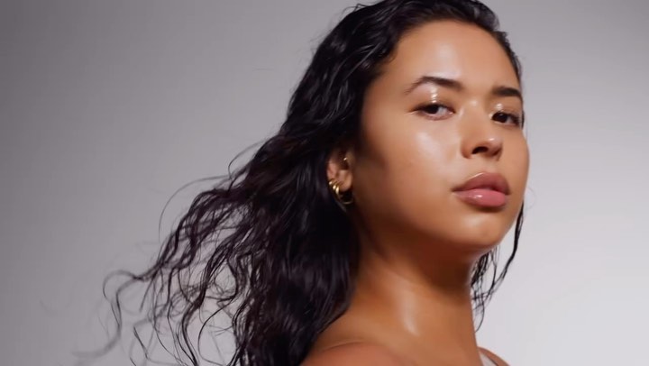 Close-up portrait of a tan-skinned woman with wet, dark, wavy hair, gold hoop earrings, and glossy makeup against a grey background.