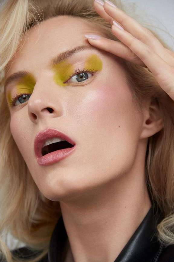 Close-up of a woman with blonde wavy hair and yellow eye shadow touching her forehead, wearing a black leather jacket against a light background.