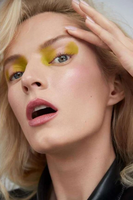 Close-up of a woman with blonde wavy hair and yellow eye shadow touching her forehead, wearing a black leather jacket against a light background.