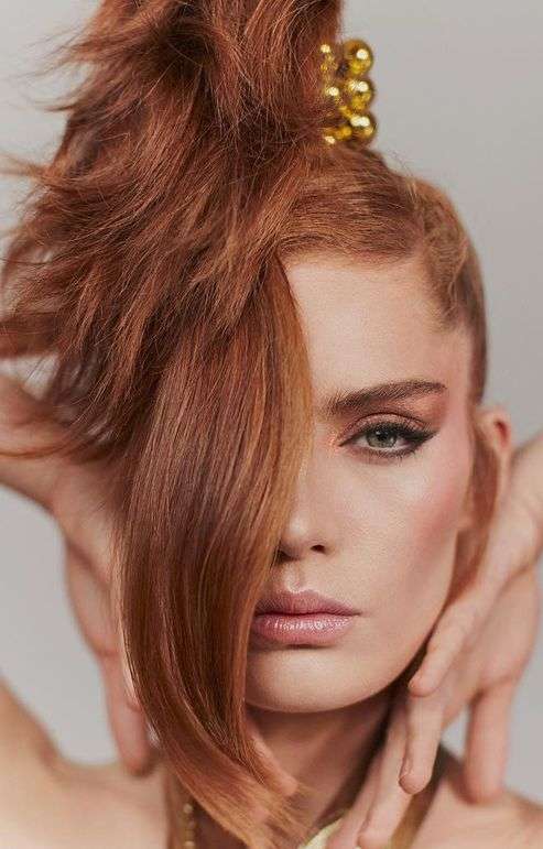 Close-up portrait of an adult female model with auburn hair styled in a high updo and styled with heavy eye makeup in a studio setting.
