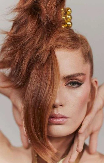 Close-up portrait of an adult female model with auburn hair styled in a high updo and styled with heavy eye makeup in a studio setting.