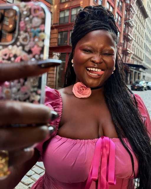 Woman in pink dress takes selfie outdoors on city street with smartphone decorated with embellishments while sticking her tongue out.