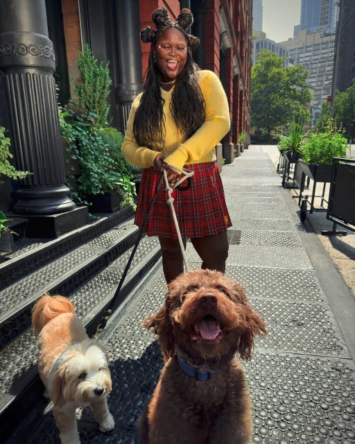 Outdoor shot of woman walking two dogs on leashes. The woman wears a yellow sweater, plaid skirt, and has a unique braided hairstyle.