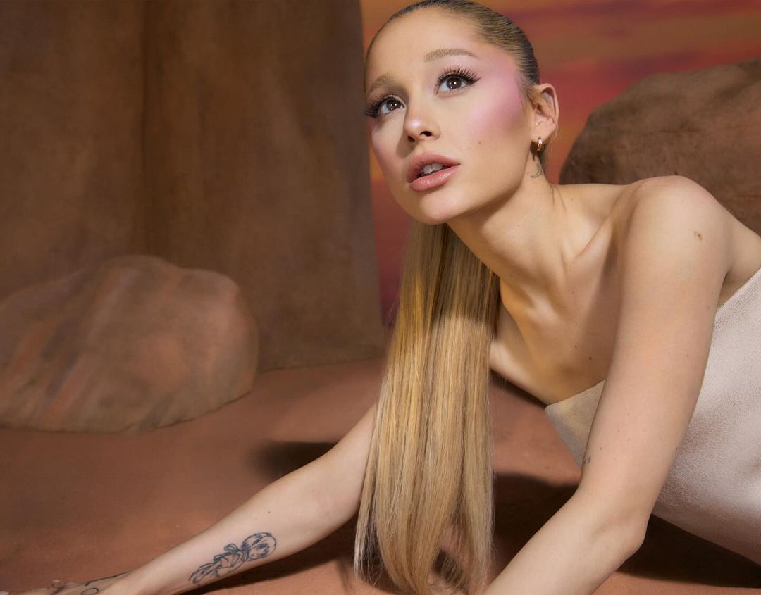 Close-up portrait of Ariana Grande with long blonde ponytail, soft pink blush, and a strapless top against a warm-toned backdrop.