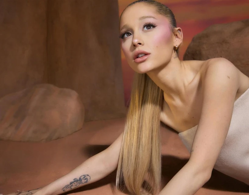Close-up portrait of Ariana Grande with long blonde ponytail, soft pink blush, and a strapless top against a warm-toned backdrop.