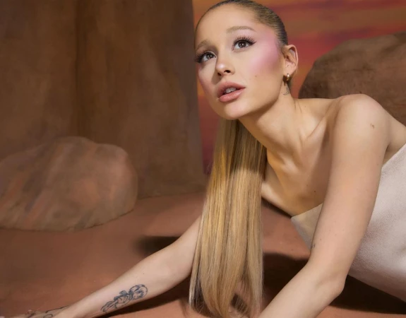 Close-up portrait of Ariana Grande with long blonde ponytail, soft pink blush, and a strapless top against a warm-toned backdrop.