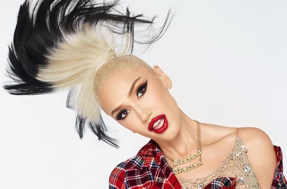 Blonde-haired woman with dramatic black wing eyeliner and red lips. She is wearing a red plaid shirt with a gold nameplate necklace.