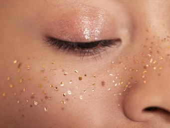 Close-up macro portrait of closed eye with glossy eyeshadow and gold leaf flakes on cheeks by Sarah Silver
