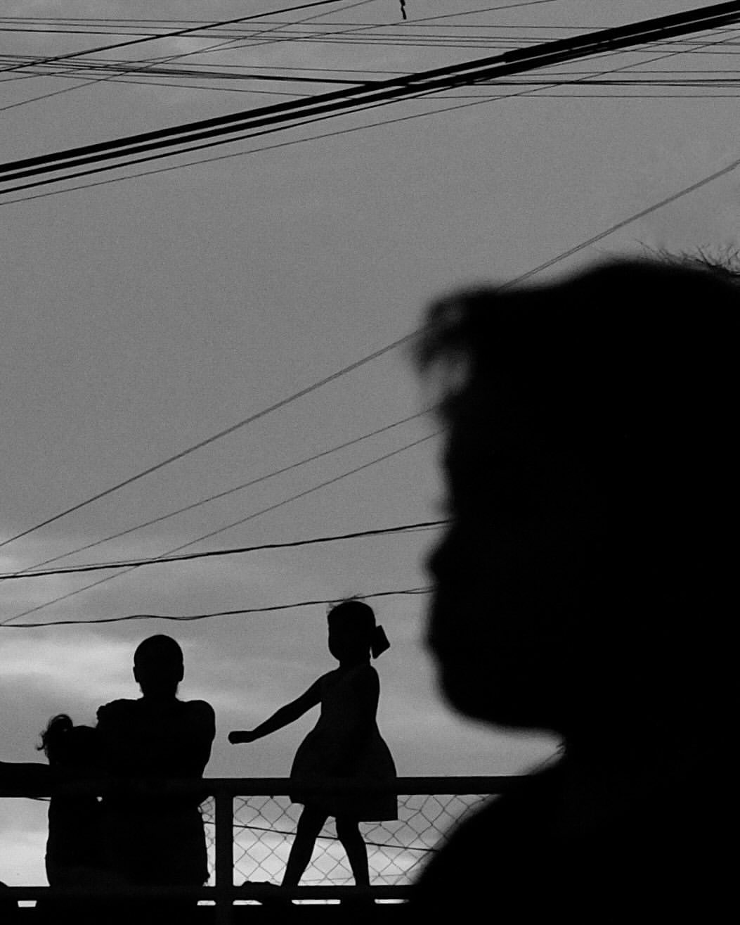 Silhouetted children on a balcony with overhead wires against a gray sky create a stark, monochrome image emphasizing shape and form.