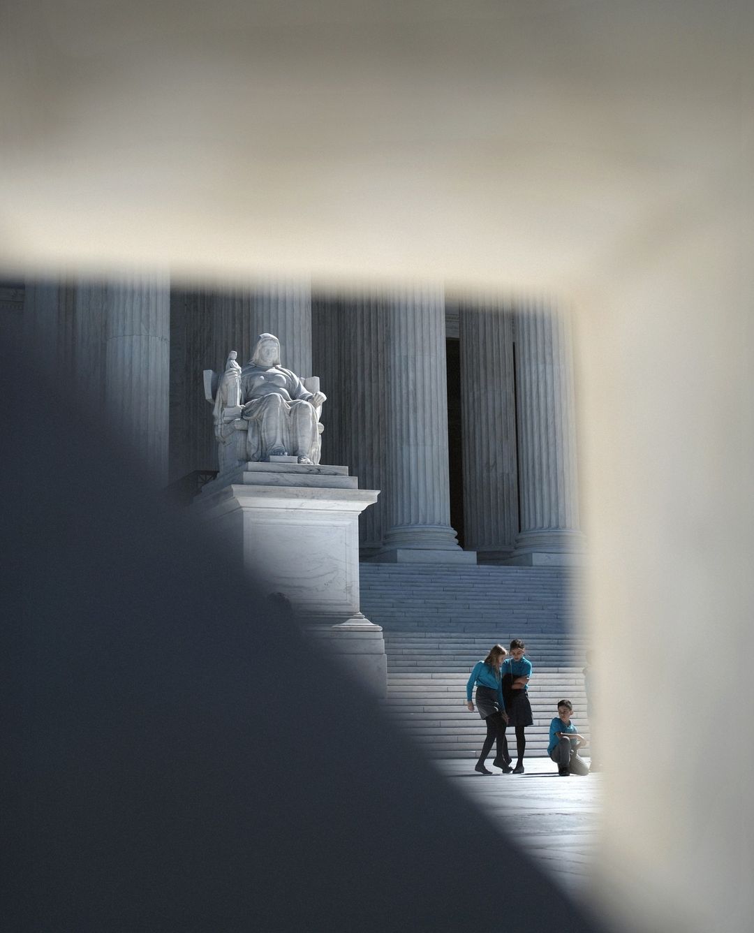 Statue of Justice stands before US Supreme Court, people gather on stairs. Sunlight highlights architecture and marble.
