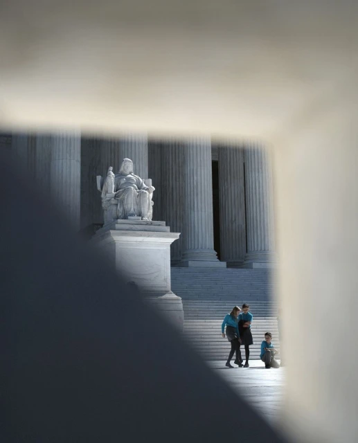 Statue of Justice stands before US Supreme Court, people gather on stairs. Sunlight highlights architecture and marble.