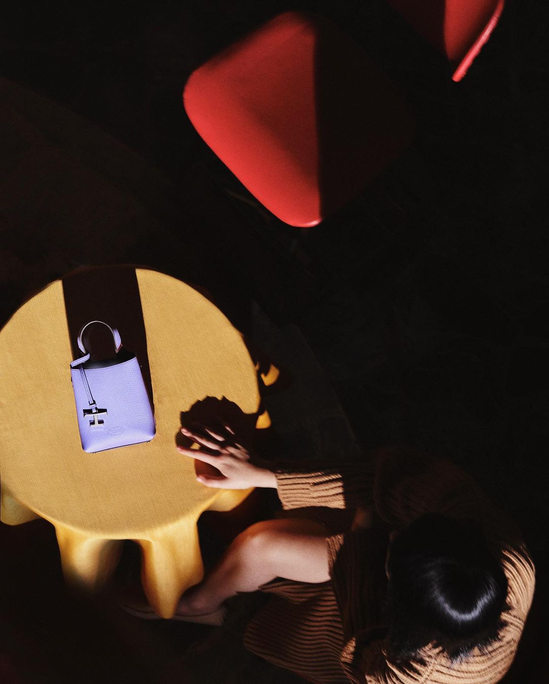 Overhead shot of a woman in a striped shirt seated at a round yellow table with a purple handbag, illuminated by dark shadows.
