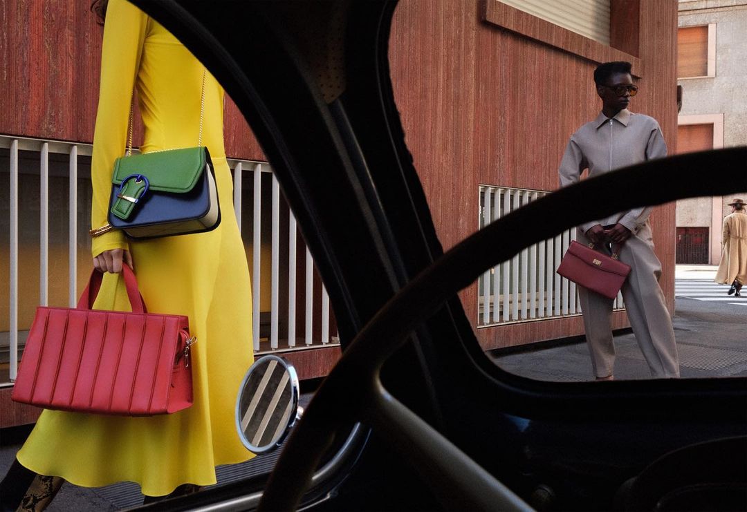 Fashion photography featuring two models in an urban setting with purses, shot from inside a car looking out the window.