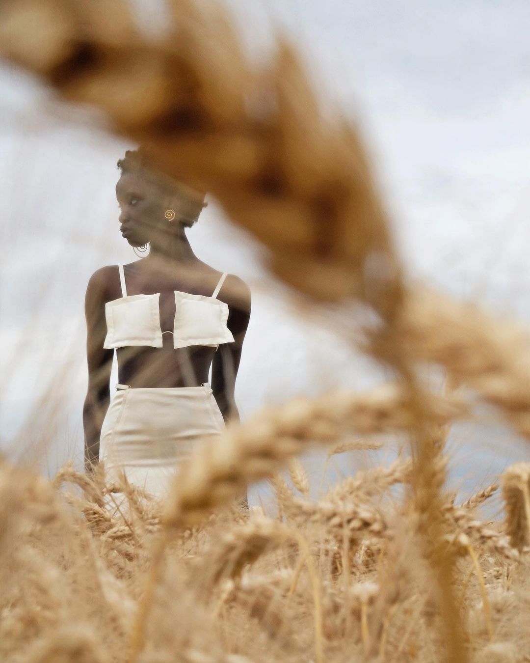 A young woman in a light-colored outfit stands in a wheat field under a cloudy sky, with some wheat stalks blurred in the foreground.