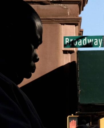 Close-up of an African American adult male in shadow, street-level view of a green Broadway sign in an urban environment, city building.