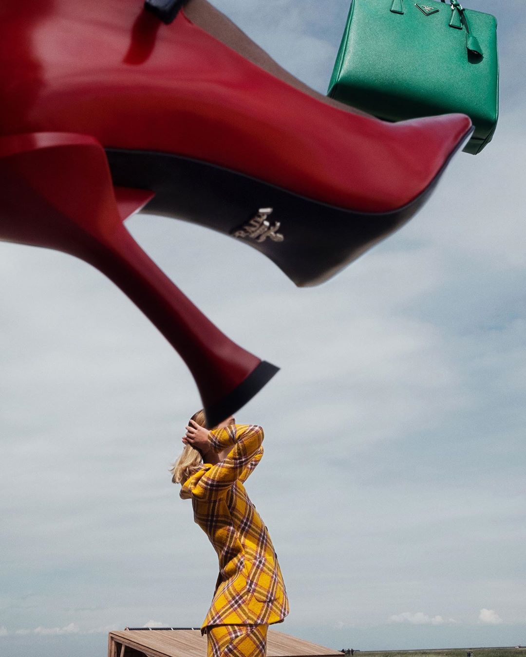 Fashion editorial photo of a woman in a yellow plaid suit looking up at a giant red high heel shoe and green handbag in a sky background.