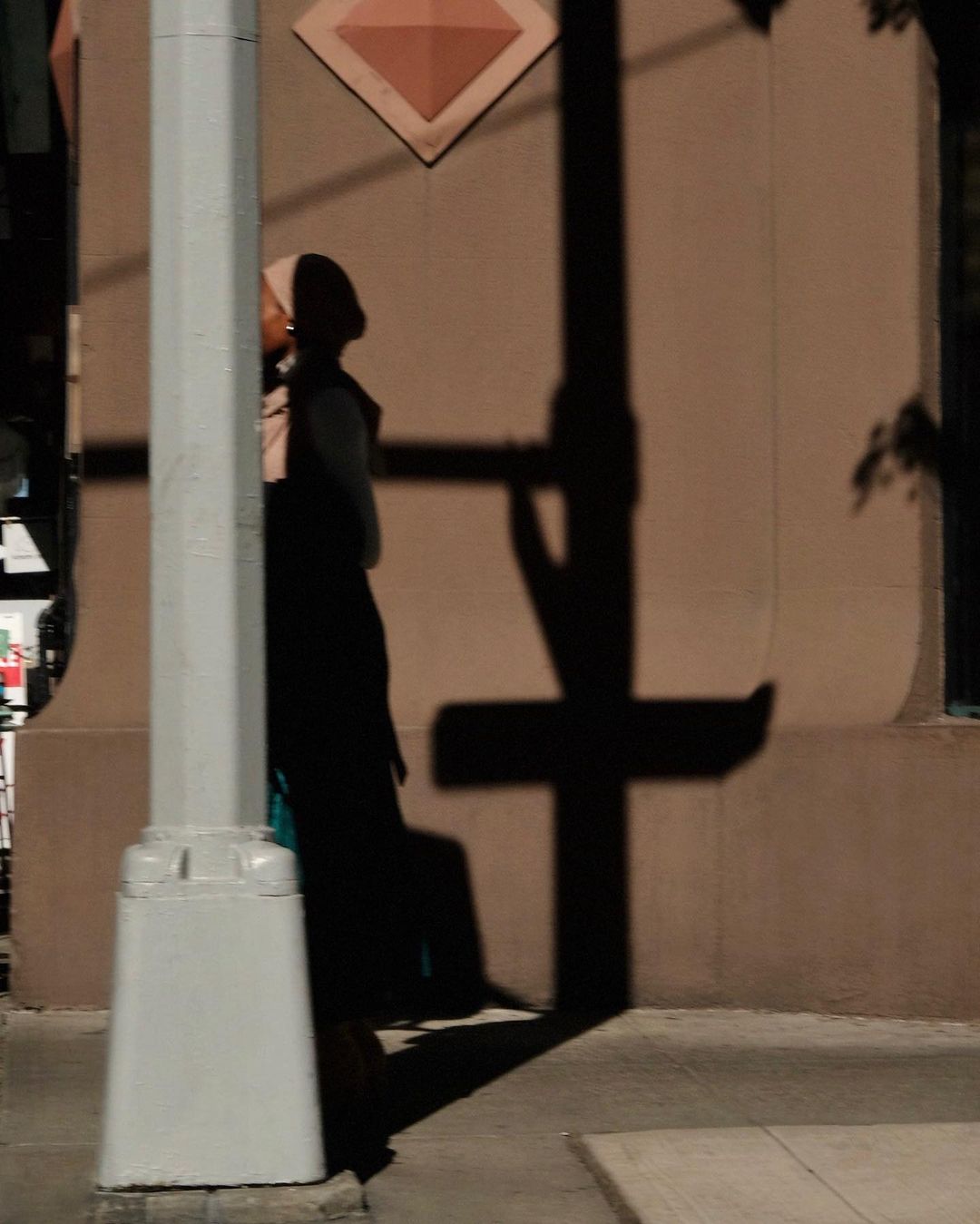 Woman in hijab stands by a white pole at dusk, casting a long shadow on a beige building exterior with diamond-shaped decoration.