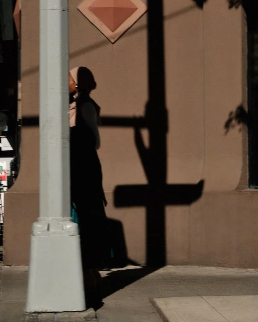 Woman in hijab stands by a white pole at dusk, casting a long shadow on a beige building exterior with diamond-shaped decoration.