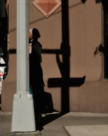 Woman in hijab stands by a white pole at dusk, casting a long shadow on a beige building exterior with diamond-shaped decoration.