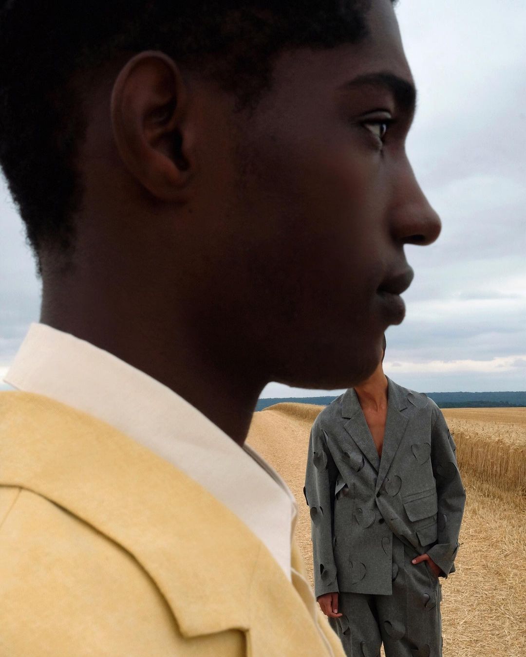 Two dark-skinned men wearing fashionable jackets. One is in focus, and the other stands at a distance on a farm landscape. 