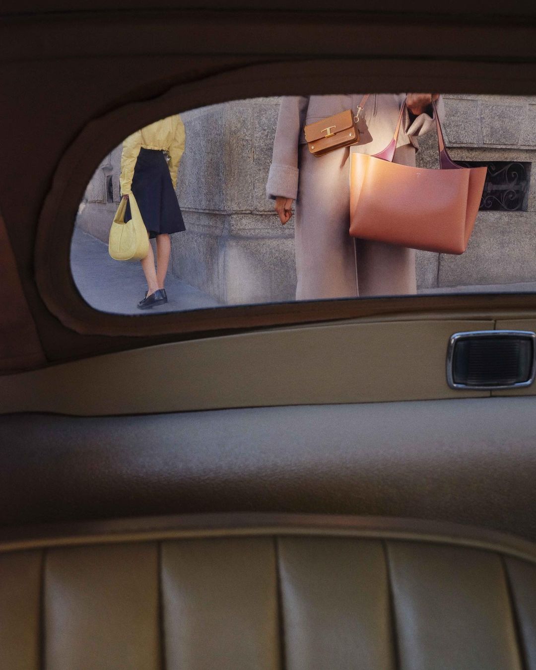 Two women stand near a building, one wearing a yellow top and navy skirt, the other in a tan coat, bags in hand, viewed from inside a car.