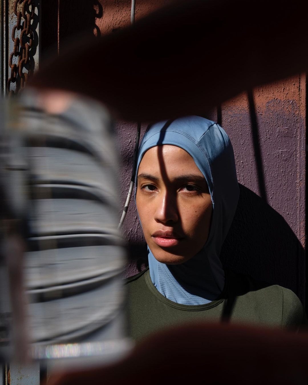 Portrait of a young adult woman wearing a blue hijab, lit by sunlight, with a partially obscured chain link fence in the background.