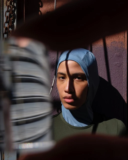 Portrait of a young adult woman wearing a blue hijab, lit by sunlight, with a partially obscured chain link fence in the background.
