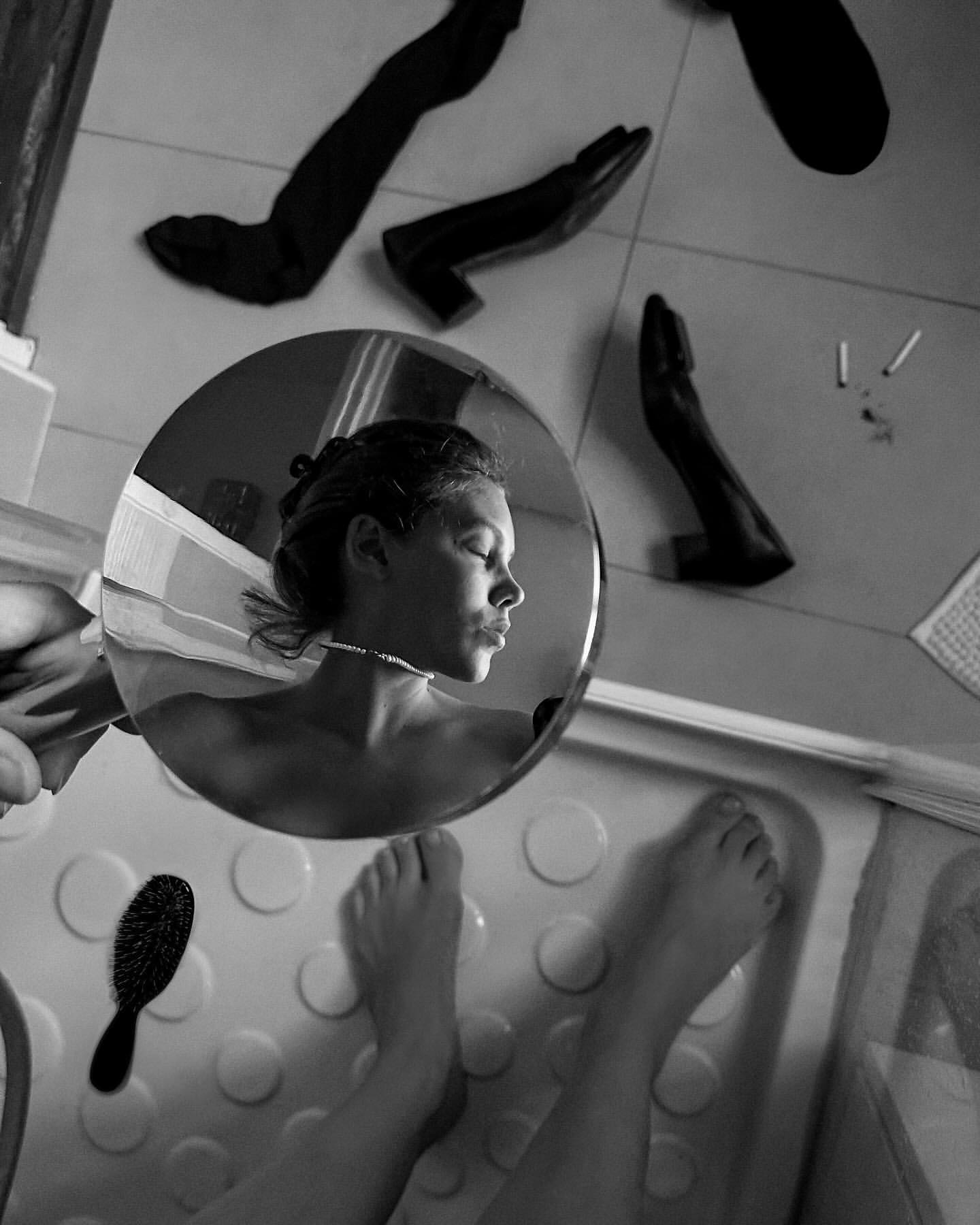 Black and white photo of a young woman in a bathtub reflected in a mirror, with floating black shoes and a hairbrush in the scene.
