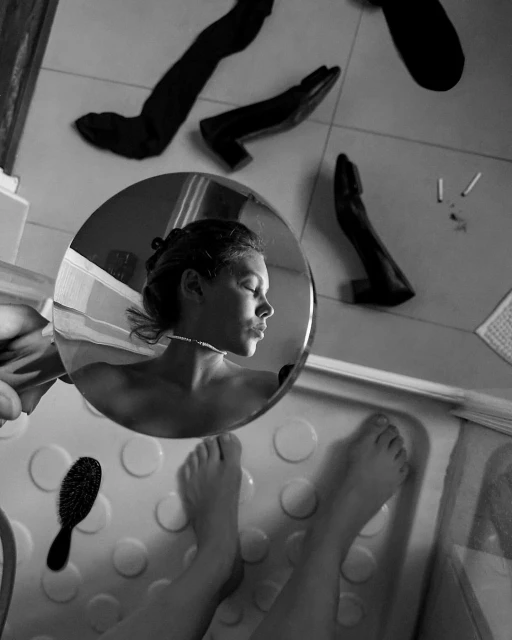 Black and white photo of a young woman in a bathtub reflected in a mirror, with floating black shoes and a hairbrush in the scene.