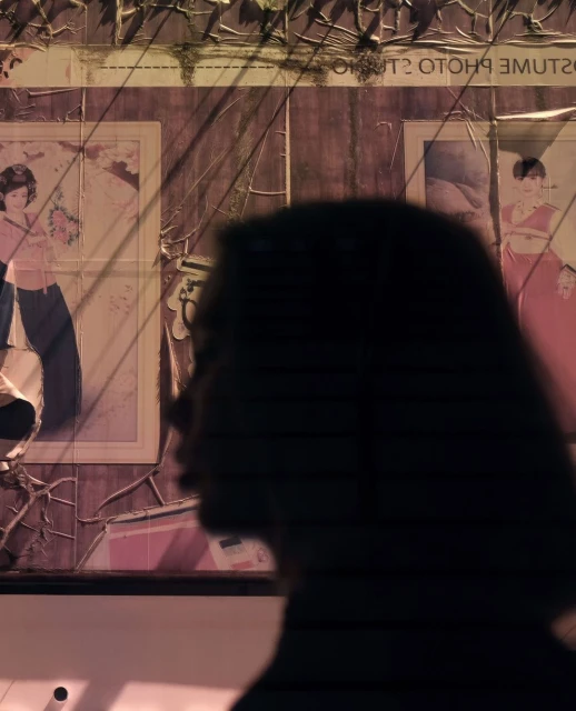 Silhouette of a person in front of vintage portrait posters on a distressed wall in an indoor setting with moody lighting.