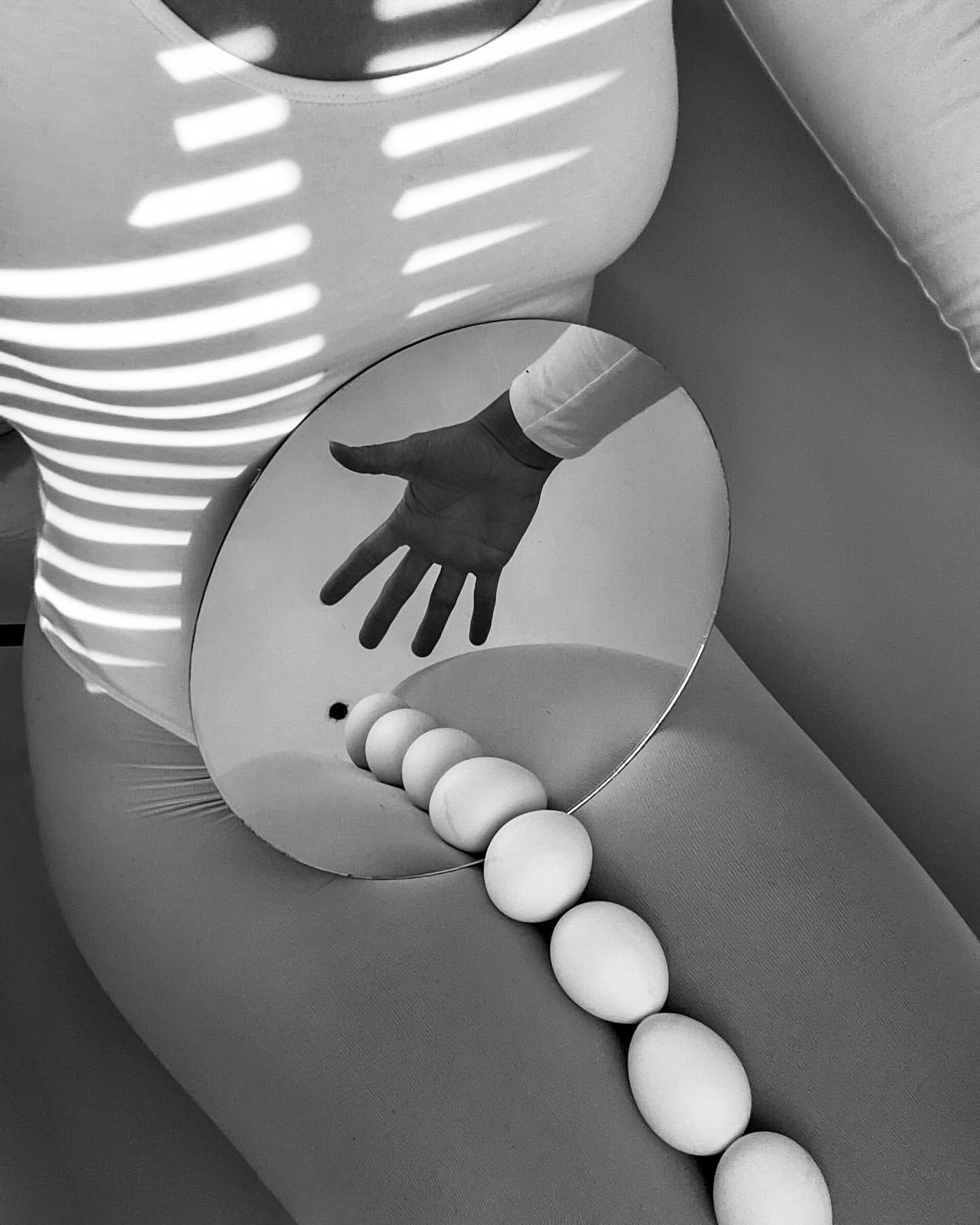 Conceptual monochrome art composition of a person with eggs and mirror creating reflections and shadows, in an indoor setting.